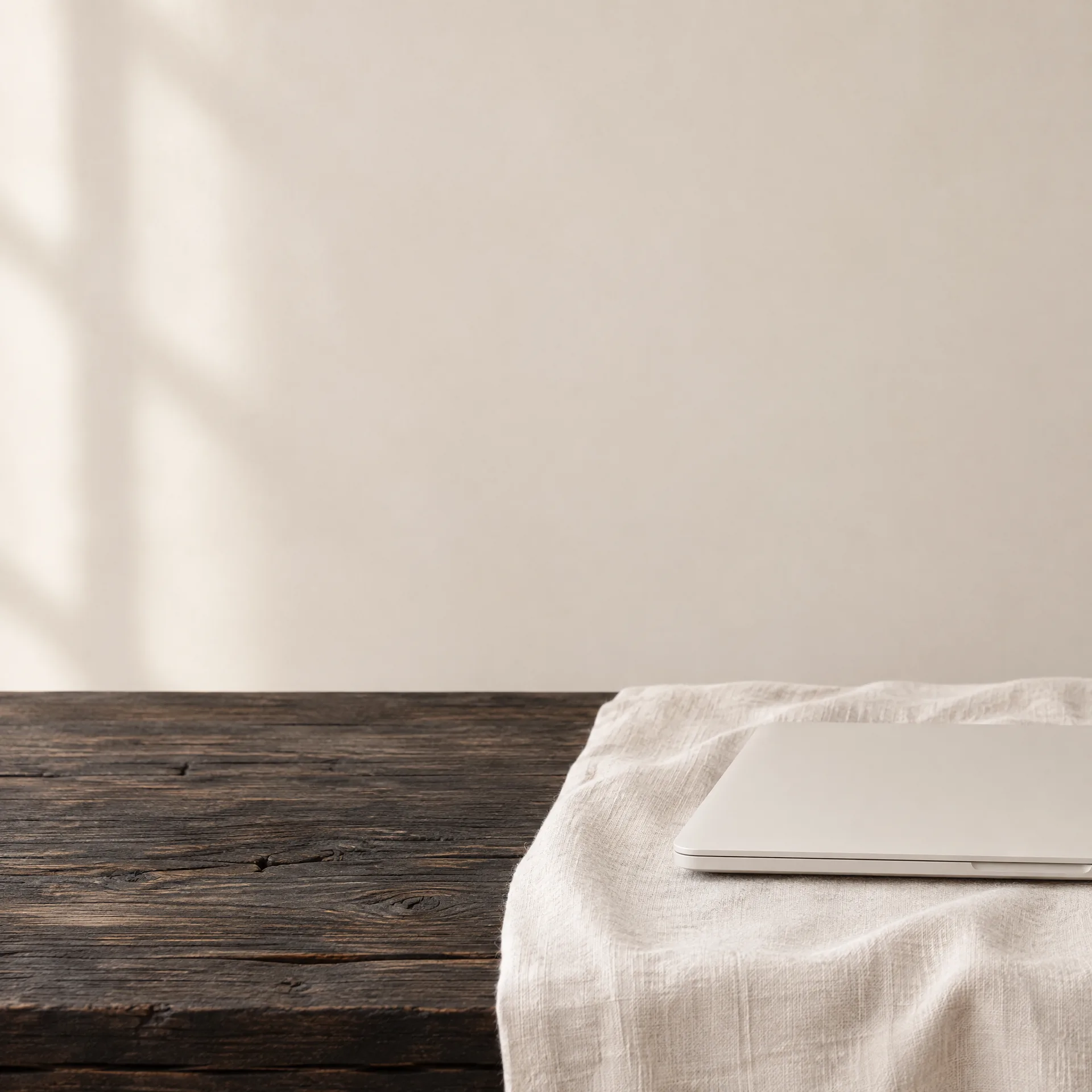 Un ordenador portátil claro y cerrado, sobre un paño de lino natural, en el borde de una mesa de madera oscura.